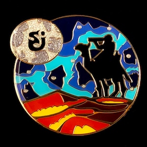 May include: A round enamel pin with a silhouette of a person riding a camel against a blue and turquoise starry night sky. The pin features a full moon with a sparkly texture and the letter "S" in the center.