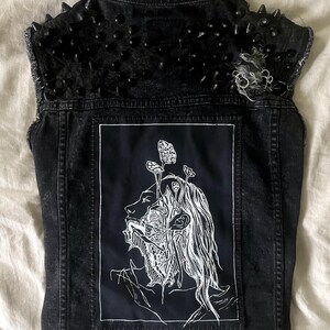 May include: Black denim vest with a distressed finish, featuring black spikes on the shoulders. The back displays a black patch with a white illustration of a face with mushroom-like elements.