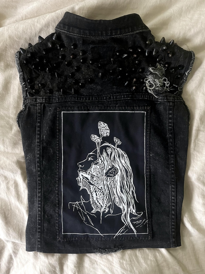 May include: Black denim vest with a distressed finish, featuring black spikes on the shoulders. The back displays a black patch with a white illustration of a face with mushroom-like elements.