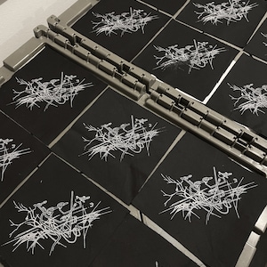 May include: A close-up of several black fabric patches with a white design of a tree with branches and birds. The patches are arranged in rows on a gray surface.
