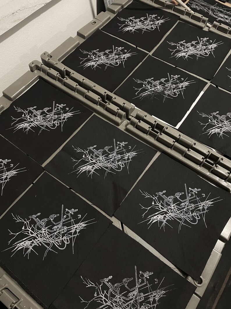 May include: A close-up of several black fabric patches with a white design of a tree with branches and birds. The patches are arranged in rows on a gray surface.