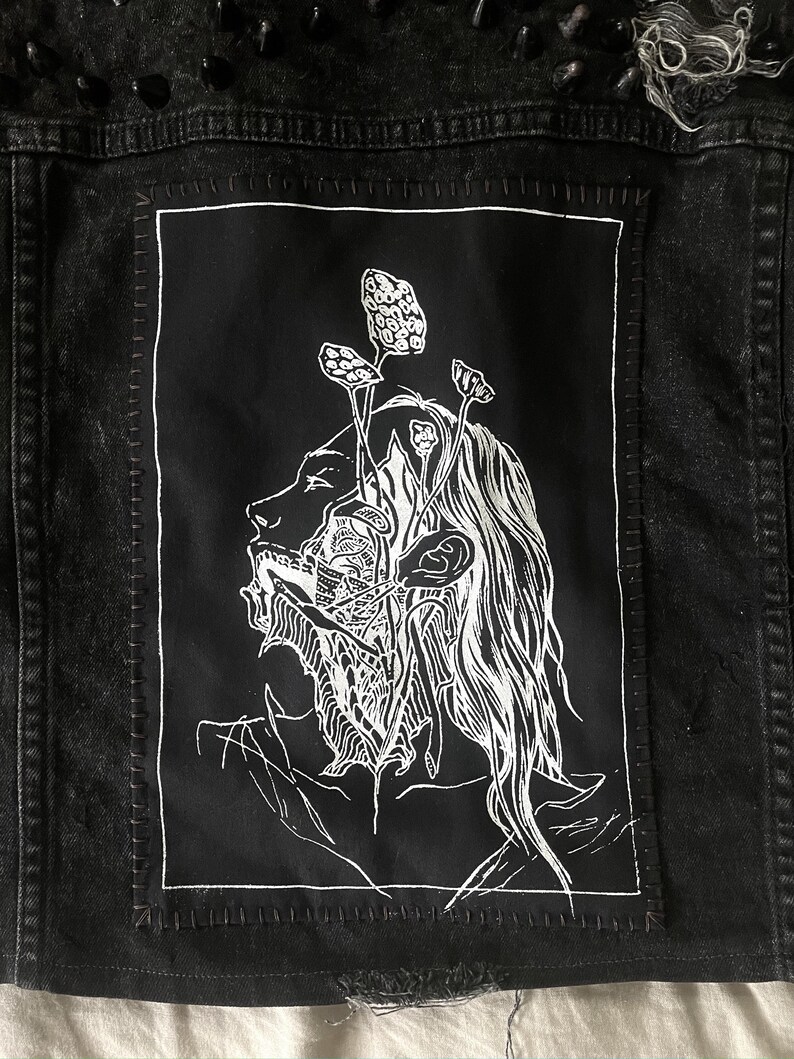 May include: Black denim jacket with a white graphic patch depicting a face with mushroom-like growths. The jacket features black studs on the shoulders and distressed detailing.