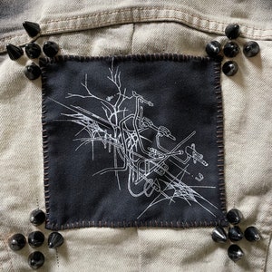May include: A black denim jacket with a black patch sewn on the back. The patch features a white graphic design of a motorcycle with a spider web. The jacket is decorated with black metal studs.