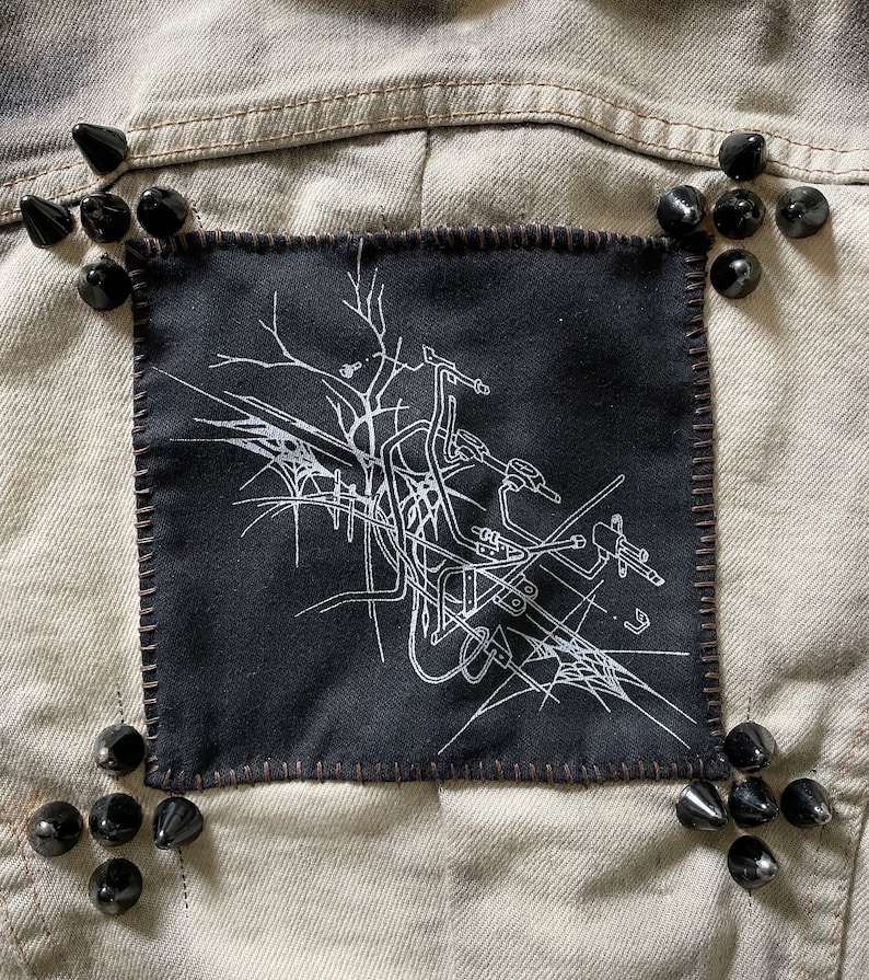 May include: A black denim jacket with a black patch sewn on the back. The patch features a white graphic design of a motorcycle with a spider web. The jacket is decorated with black metal studs.