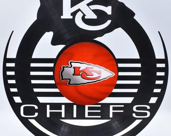 May include: A black vinyl record with a white outline of a Native American headdress and the text "KC" inside. The centre of the record has a red and white Kansas City Chiefs logo. The text "CHIEFS" is printed in white below the logo.