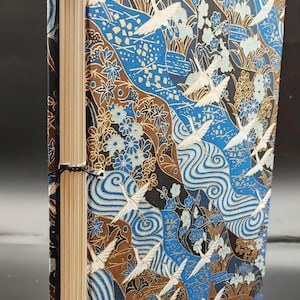 May include: A handmade journal with a blue, brown and gold patterned cover featuring white cranes flying over a river. The cover is made of Japanese paper and the journal is bound with black thread.