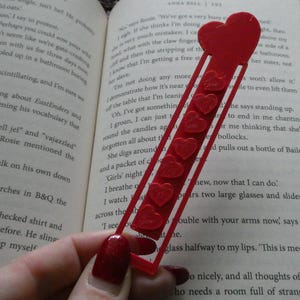 May include: A red heart-shaped bookmark with six small heart details. The bookmark is made of a thin, transparent material and is in an open book. A decorative accessory for book lovers.