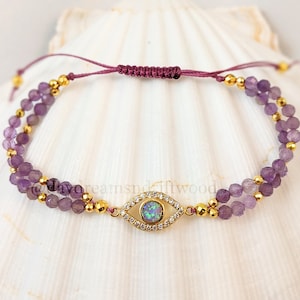 May include: A double-strand bracelet featuring a gold evil eye charm. The bracelet is made with purple beads and gold beads. The evil eye charm has a clear stone in the centre and is surrounded by smaller clear stones.