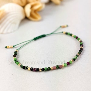 May include: A green, black, pink and gold beaded bracelet with a green cord and a sliding knot fastening. The bracelet is made with small, round beads.