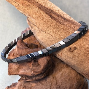 Op de afbeelding: Een zwarte hematiet armband met een geometrisch ontwerp. De armband is gemaakt van kleine, vierkante kralen die met elkaar verbonden zijn om een ​​continue ketting te vormen. De armband ligt op een stuk drijfhout.