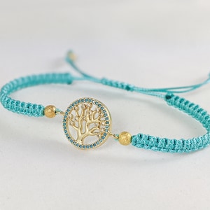May include: A turquoise braided bracelet with a gold tree of life charm. The charm is surrounded by small turquoise stones.