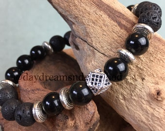 May include: Black lava rock and onyx bead bracelet with a silver charm. The charm has a geometric design with black accents.