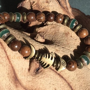K&ouml;nnte beinhalten: Ein braunes Holzperlenarmband mit gr&uuml;nen und goldenen Akzenten. Das Armband hat einen Fisch-Anh&auml;nger.