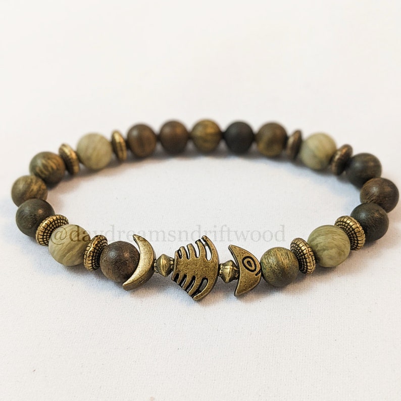 K&ouml;nnte beinhalten: Ein Holzperlenarmband mit einem Metallfisch-Anh&auml;nger. Die Perlen sind in Braun- und Gr&uuml;nt&ouml;nen.