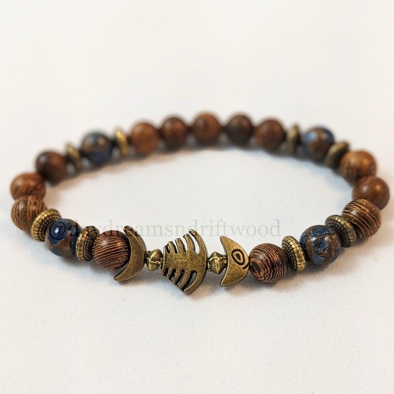 K&ouml;nnte beinhalten: Ein braunes Holzperlenarmband mit einer blau-braunen Perle und einem Fisch-Anh&auml;nger aus Metall.