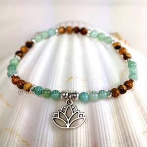 May include: A green and brown beaded bracelet with a silver lotus charm. The bracelet is on a white seashell background.