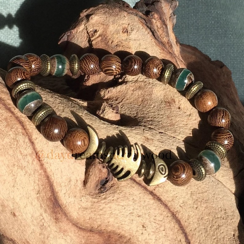 K&ouml;nnte beinhalten: Ein braunes Holzperlenarmband mit gr&uuml;nen und goldenen Akzenten. Das Armband hat einen Fisch-Anh&auml;nger.