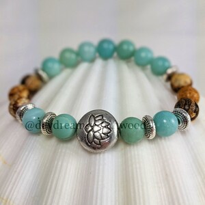May include: A turquoise and brown beaded bracelet with a silver charm featuring a lotus flower design. The bracelet is on a white seashell background.