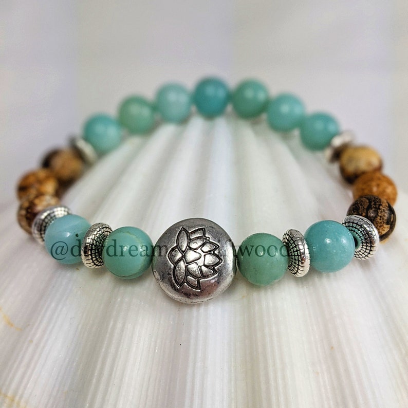 May include: A turquoise and brown beaded bracelet with a silver charm featuring a lotus flower design. The bracelet is on a white seashell background.