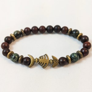 K&ouml;nnte beinhalten: Ein braunes und gr&uuml;nes Perlenarmband mit einem goldenen Fischgr&auml;ten-Anh&auml;nger. Das Armband besteht aus nat&uuml;rlichen Steinen und hat einen rustikalen Look.