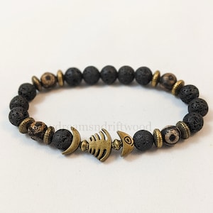 K&ouml;nnte beinhalten: Schwarzes Lava-Armband mit einem goldenen Fisch-Anh&auml;nger. Das Armband besteht aus kleinen, runden Perlen und hat ein schlichtes, elegantes Design.