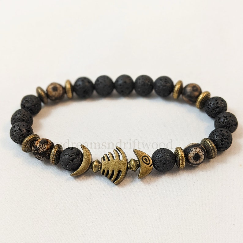 K&ouml;nnte beinhalten: Schwarzes Lava-Armband mit einem goldenen Fisch-Anh&auml;nger. Das Armband besteht aus kleinen, runden Perlen und hat ein schlichtes, elegantes Design.
