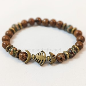 K&ouml;nnte beinhalten: Ein braunes Holzperlenarmband mit einem Metallfisch-Anh&auml;nger. Die Perlen sind eine Mischung aus Braun und Gr&uuml;n.