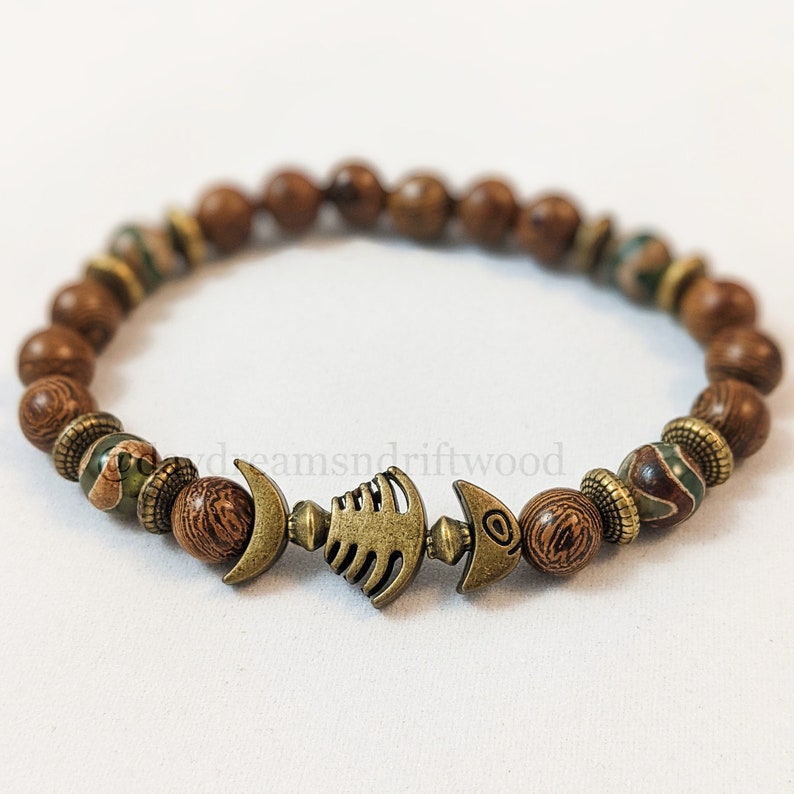 K&ouml;nnte beinhalten: Ein braunes Holzperlenarmband mit einem Metallfisch-Anh&auml;nger. Die Perlen sind eine Mischung aus Braun und Gr&uuml;n.