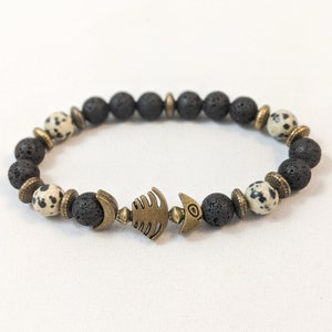 K&ouml;nnte beinhalten: Schwarzes und wei&szlig;es Lava-Armband mit einem bronzenen Fisch-Anh&auml;nger. Das Armband besteht aus nat&uuml;rlichen Steinen und ist ein tolles Accessoire f&uuml;r jedes Outfit.