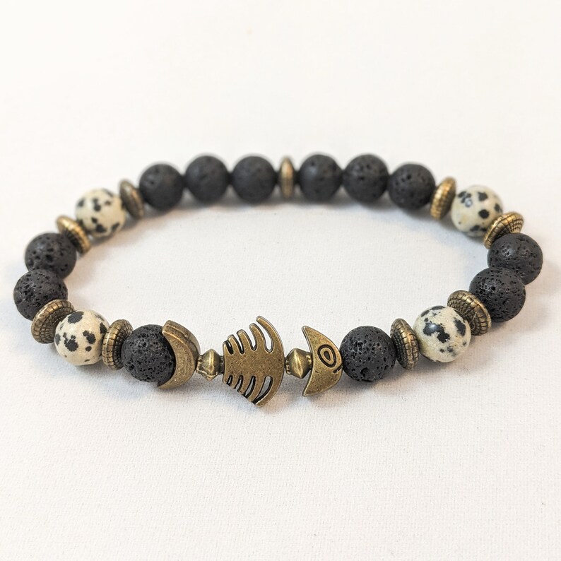K&ouml;nnte beinhalten: Schwarzes und wei&szlig;es Lava-Armband mit einem bronzenen Fisch-Anh&auml;nger. Das Armband besteht aus nat&uuml;rlichen Steinen und ist ein tolles Accessoire f&uuml;r jedes Outfit.