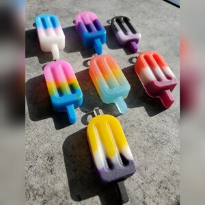 Könnte beinhalten: Ein Set aus zehn bunten Eis am Stiel-Anhängern mit verschiedenen Farbkombinationen, darunter Blau, Rosa, Lila, Gelb und Weiß. Die Anhänger sind aus Harz gefertigt und haben eine glänzende Oberfläche.