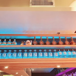 May include: A wooden shelf with two rows of liquor bottles. The top row has brown bottles with gold labels. The bottom row has clear bottles with blue labels. The bottles are arranged in a symmetrical pattern.