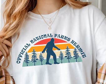 Pode incluir: Camiseta branca com um design retrô com uma silhueta de Bigfoot, árvores e um pôr do sol. O texto "OFFICIAL NATIONAL PARKS HERMIT" está em arco acima do gráfico. A palavra "WHITE" está impressa na parte inferior.