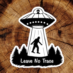 Peut inclure: Autocollant noir et blanc représentant un OVNI en train d'enlever une silhouette de Bigfoot. Le design comprend une silhouette de forêt et le texte "Leave No Trace".