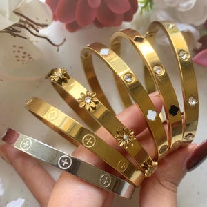 May include: A collection of gold and silver bangles. Some bangles feature flower and gemstone accents, while others have cross symbols. The bracelets are displayed against a backdrop of flowers.