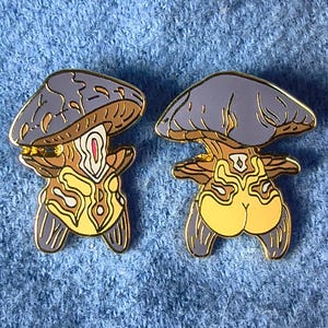 Puede incluir: Dos pines de esmalte con forma de hongo. Cada pin presenta una tapa azul, un tallo marrón y un cuerpo amarillo con intrincados detalles dorados. Los pines tienen un contorno dorado y están sobre un fondo azul texturizado.