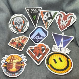 Peut inclure: Un ensemble de neuf autocollants en vinyle présentant divers motifs graphiques, dont un crâne, un visage souriant jaune, un champignon atomique avec le texte Droz & Davis The Last Resort, et des logos avec Vinson Dynamics, Aces et IMC.