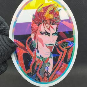 May include: Oval sticker featuring a stylized illustration of a person with red hair and makeup, sticking out their tongue. The sticker has a holographic effect and includes the non-binary pride flag colors in the background.