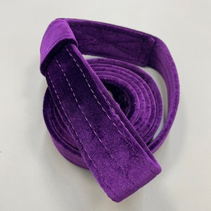 May include: A deep purple velvet strap coiled into a circular shape. The strap features a soft, plush texture with visible double-stitched detailing running along the length of the fabric, creating a structured and durable appearance for the material.