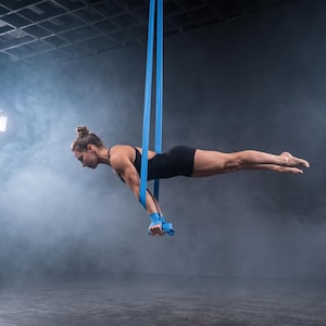 May include: A pair of bright blue aerial silk straps suspended from above, featuring durable fabric loops used for suspension training and acrobatics. The straps are solid blue, made of a smooth, flexible material designed to support body weight.