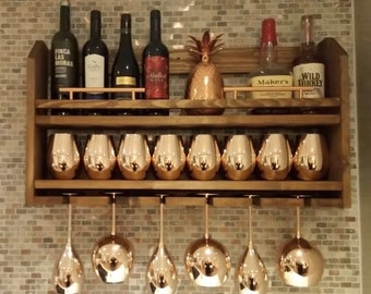 May include: A wooden wall-mounted wine rack with two shelves. The top shelf holds five bottles of wine and two bottles of liquor. The bottom shelf holds eight copper-colored wine glasses. The rack has a copper-colored metal bar across the front of each shelf.