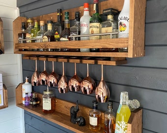 Könnte beinhalten: Ein wandmontiertes Barregal aus Holz mit zwei Ebenen. Die obere Ebene hat ein Metallgestell für Gläser und ist mit verschiedenen Flaschen Alkohol gefüllt. Die untere Ebene hat ein Regal für Flaschen und Gläser und verfügt über zwei kleine Elefantenfiguren. Das Regal ist in einem dunklen Braunton gebeizt.