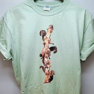 May include: A light green t-shirt with a graphic print of a stack of squirrels climbing a tree trunk.