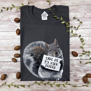 May include: A dark gray t-shirt with a graphic of a squirrel holding a sign that says "Take me to your feeder".