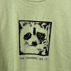 May include: Light green t-shirt featuring a black and white raccoon graphic inside a square frame. The raccoon has a surprised expression and is holding onto a wooden post. The text "THE SQUIRREL DID IT..." is printed below the graphic.