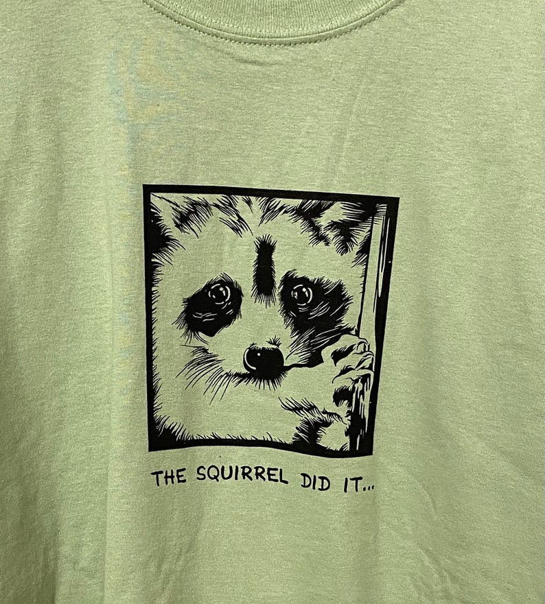 May include: Light green t-shirt featuring a black and white raccoon graphic inside a square frame. The raccoon has a surprised expression and is holding onto a wooden post. The text "THE SQUIRREL DID IT..." is printed below the graphic.