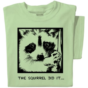 May include: Light green t-shirt featuring a black and white illustration of a raccoon inside a black square. The raccoon is peeking out, with the text "THE SQUIRREL DID IT..." below the image. The shirt is folded.