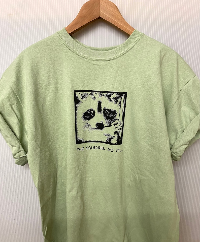 May include: A light green t-shirt with a black and white raccoon graphic inside a black square. The text "THE SQUIRREL DID IT..." is below the graphic. The shirt is hanging on a wooden hanger.