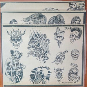 Op de afbeelding: Een vel met zwart-witte tattoo-flash met verschillende schedelontwerpen, waaronder een piraten schedel, een slangen schedel en een demonen schedel. De flash is gelabeld met "B-R-A" en "B-R-B".