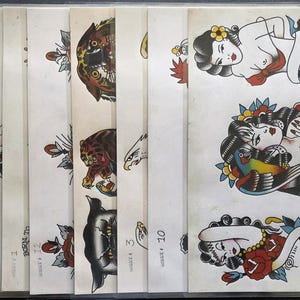 Può includere: Una collezione di dieci fogli di carta color avorio con disegni di tatuaggi tradizionali. Le illustrazioni includono tigri, aquile e varie figure femminili con fiori, realizzate in uno stile grafico audace, colorato e classico.